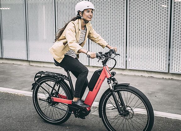 Elektroradtypen Eine Frau fährt auf einem roten Elektrorad auf Asphalt, im Hintergrund graue, gelochte Metallwände.