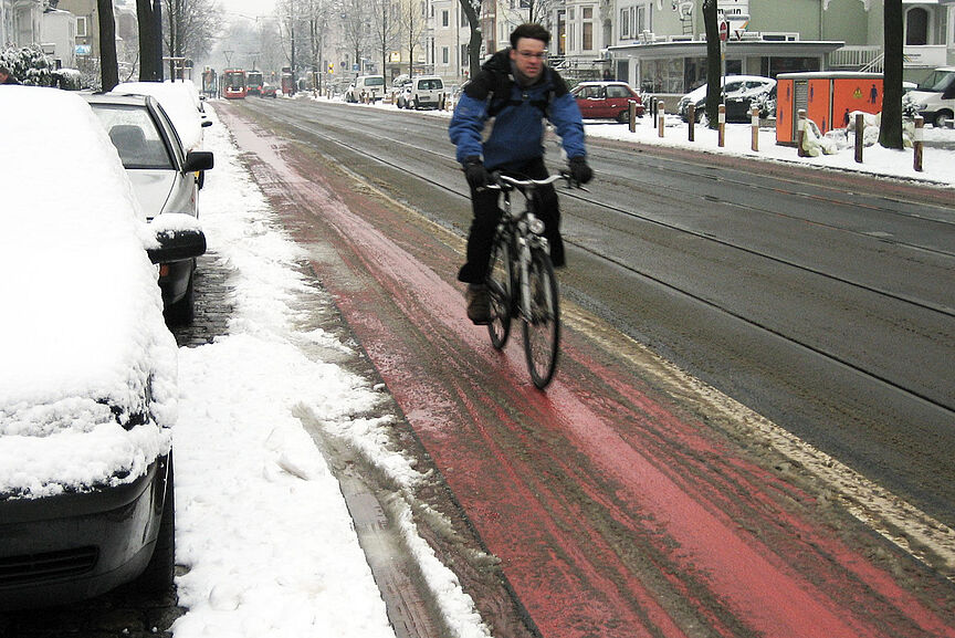 Radfahren im Winter Ein Radfahrer in Winterkleidung fährt auf einem rot markierten Radfahrstreifen neben einer Straße, der weitgehend von Schnee befreit ist, während am Straßenrand und auf geparkten Autos Schnee liegt.